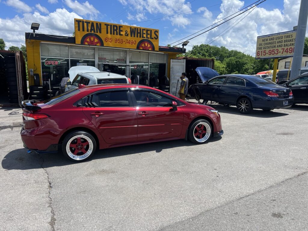Side profile of a custom Toyota Corolla parked in front of Titan Tire & Wheels shop.