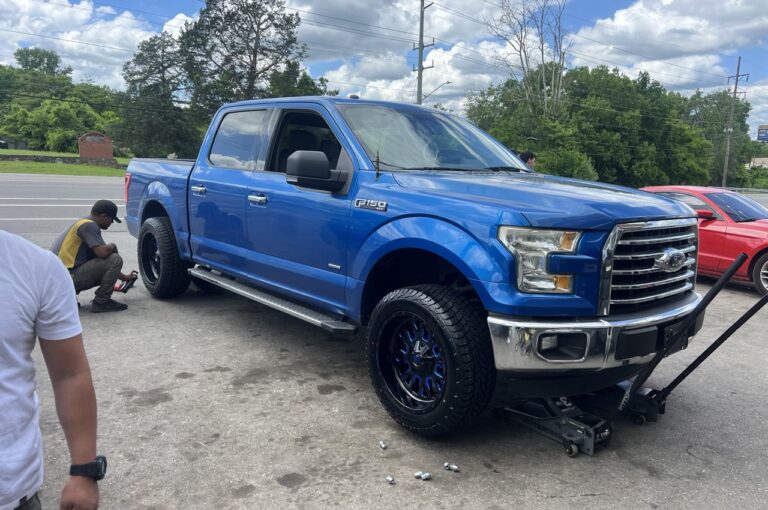 A blue Ford F-150 being serviced with new Fuel wheels and Rovelo tires by a technician outside a tire shop.