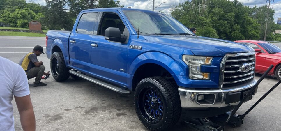 A blue Ford F-150 being serviced with new Fuel wheels and Rovelo tires by a technician outside a tire shop.